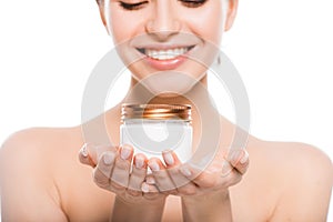 View of naked young woman holding container with face cream and smiling isolated on white