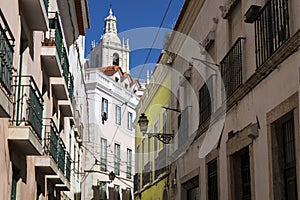 View of a narrow street and buildings with a tower of the Sao Vincente de Fora church on the backrgound, in the historic neighbour