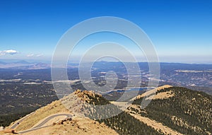 View from near top above the tree line of Pikes Peak Colorado of hairpin curve road with panorama in distance including a lake - s