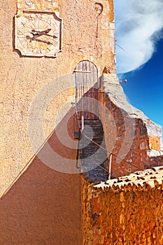 View on ochre natural stone fortress wall tower with clock, steep narrow stairs to ancient wooden entrance door, blue sky -