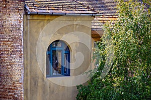 View of an old brick building with an arched window and tiled roof. The plastered facade of the house. Picturesque green tree in