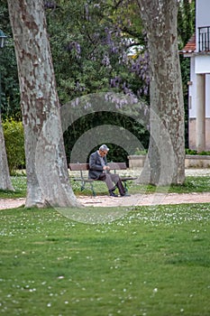 View of old man sitting on a bench, behind the garden trees, resting and looking around the park.olderTomar / Portugal - 04 04