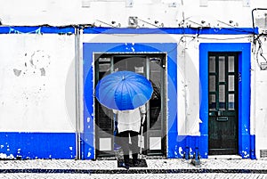 View on Old man with umbrella in front of blue colored building in the city of Ericeira, Portugal