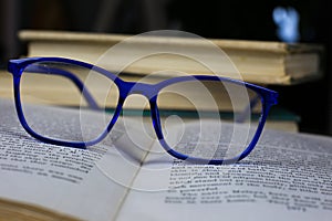 View on old open yellowed book with blue reading glasses on antique wood table with blurred pile of books background