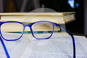 View on old open yellowed book with blue reading glasses on antique wood table with blurred pile of books background