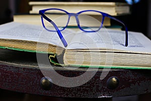 View on old open yellowed book with blue reading glasses on antique wood table with blurred pile of books background