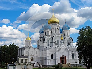 View of old Orthodox white stone church with gold and blue domes in monastery against background of green trees and cloudy sky.