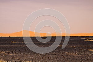 View over endless burned black flat waste stony land on golden sand dunes and blurred gloomy sky, Erg Chebbi, Morocco