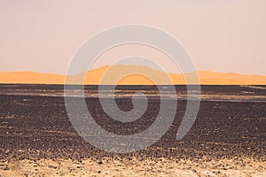 View over endless burned black flat waste stony land on golden sand dunes and blurred gloomy sky, Erg Chebbi, Morocco