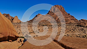 View over mountain Spitzkoppe, Kalahari, Namibia, with water pond and round shaped brownish rocks at sunny day with blue sky.