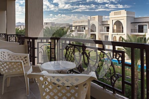 View at palm trees and a hotel foreside from a terrace with chairs and a table. Sunny day with blue sky and white clouds