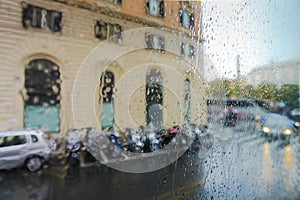 View on the parked scooters and cars on street of Rome on the rainy and day through wet glass. Italy, Rome.