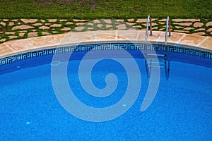 View of a part of the public pool with blue clear water: descent into the pool with perrils, sides, natural stone and grass.