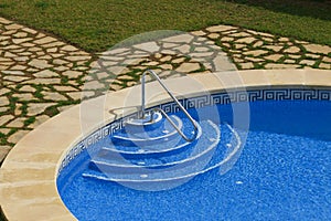 View of a part of the public pool with blue clear water: descent into the pool with perrils, sides, natural stone and grass.