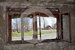 View of paved road and houses from inside of abandoned house through destroyed window with broken window frame mounted on cracked