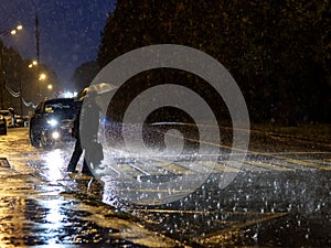 View of a pedestrian crossing in the city at night during a heavy downpour. Silhouettes of people with umbrellas.