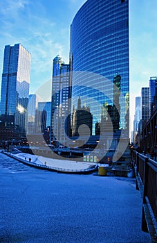 View of pedestrians on a riverwalk on a blue and frigid winter morning in Chicago alongside a frozen river and elevated bridge.