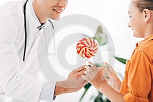 View of pediatrist in white coat giving candy to child in clinic