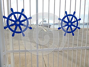 View of the port through the white entrance gates depicting two blue rudders in the background a ship docked at the port