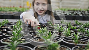 View of pretty smiling girl in greenhouse, looks and touches flower