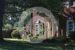 View of pretty two story brick house with bay windows and tall trees and red front door from side angle in late afternoon with hig