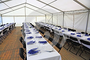 View of rented tent ready for guests. White table cloth, white plates with blue napkins and empty glass. Party celebration concept