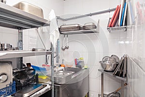 View of restaurant's professional washer, sink, brushes, metal shelving and shelves with kitchen utensils, cups, cutting board,