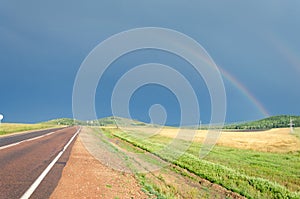 View of the road and rainbow in the sky. On the side of the road fields with green grass and forest