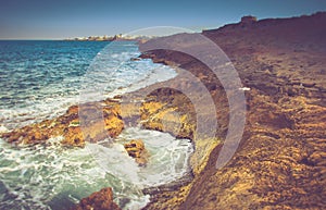 View of the rocky coast, foaming waves of the sea and the city in the distance at sunset.