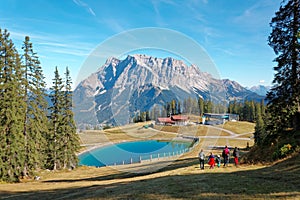 View of rugged Mountain Zugspitze on a brisk sunny day with tourists walking toward a blue pond at the foothills