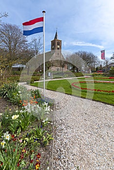 View of a rural church in a Dutch village in early spring, in the garden there are several flowers and prominently the Dutch flag,