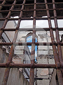 View through rusty iron bars on a window of a ruined collapsing building with crumbling walls and open to the sky