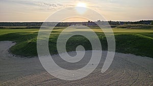 View of a sand bunker on a golf course, with green turf and the setting sun in the background.