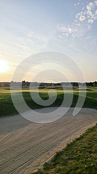 View of a sand bunker on a golf course, with green turf and the setting sun in the background.