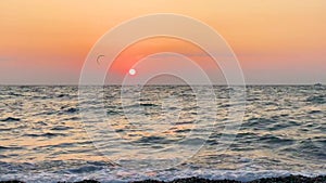 View of the sea horizon with waves on the beach, in the distance people ride kitesurfing in the evening at sunset summer time