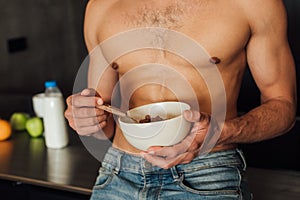 View of shirtless man holding bowl with cornflakes near fruits and bottle with milk in kitchen