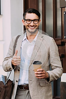 View of smiling businessman in glasses holding coffee to go and showing thumb up on street