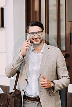 View of smiling businessman with leather bag talking on smartphone and showing thumb up on street