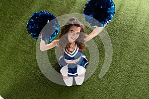 View of smiling cheerleader girl in blue uniform sitting with pompoms on green field