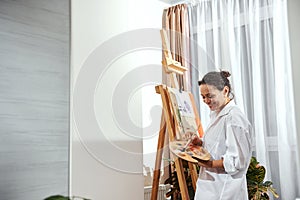 View of a smiling female painter with gathered hair in a bun and paint brushes in her hair standing in front of the easel in