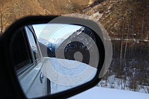 View of the snow-capped mountains and the winter road in the rearview mirror of the car
