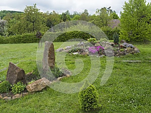 View on spring garden with oval flower bed with green herbs with big sandstone rock and blooming rock garden on