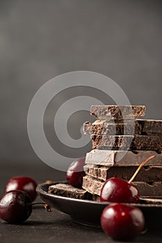 View of stack of chocolate bars in plate on wooden table with cherries, selective focus, gray background,