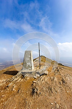 A view of the summit trig point of a Scottish mountain Ben Vorlich with eroded rocky soil under a majestic blue sky and altitude