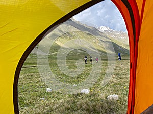 View from the tent: climbers with backpacks and trekking poles walk through alpine meadows
