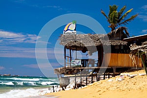 View on terrace of wooden beach bar with palm tree, strong waves, sand beach against blue sky on windy day