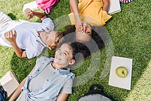 View of three happy multicultural schoolkids smiling while lying on lawn