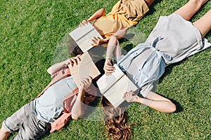 View of three multicultural schoolkids lying on lawn and covering faces with books