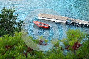 View to calm sea bay from above through some coniferous trees, red rescue boat docked near small wooden pier