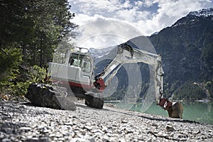View to small excavator on gravel shore at alp lake with forest